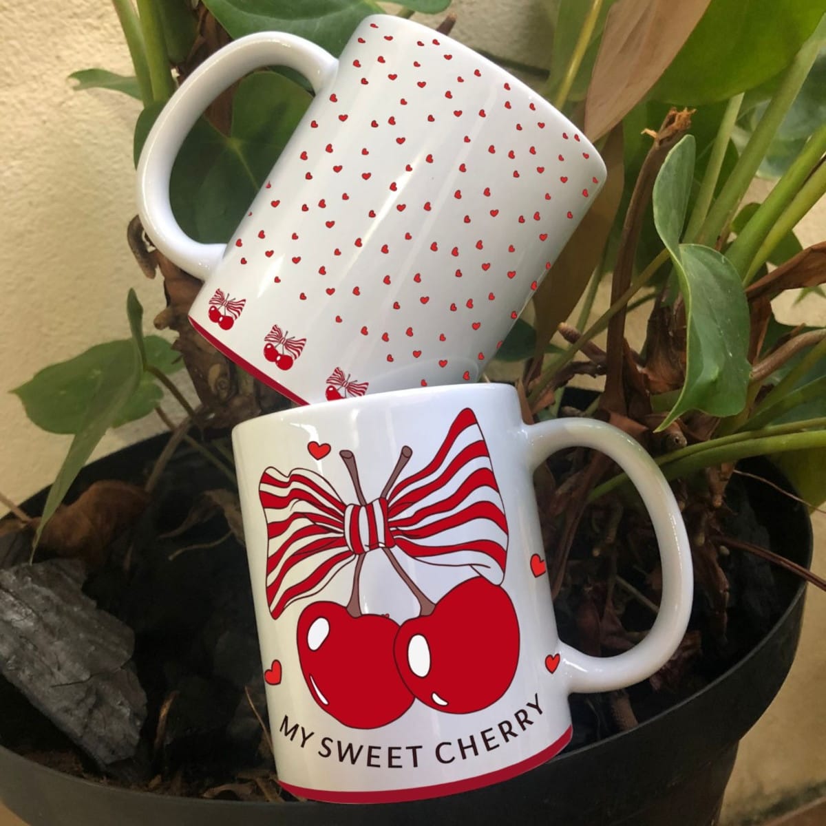 Arte Digital Caneca Cerejas PNG e Corel para Sublimação 2 Estampas de cerejas, laços e corações decoram canecas brancas em arranjo com planta.