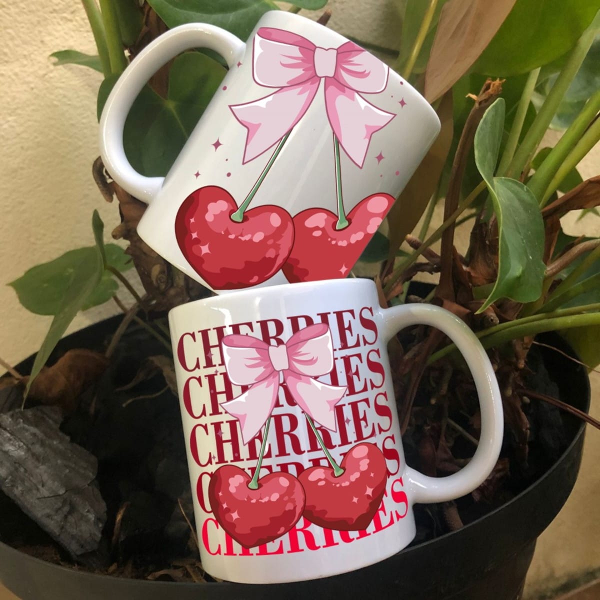 Arte Digital Caneca Cerejas PNG e Corel para Sublimação 3 Duas canecas brancas com estampa de laço rosa e cerejas vermelhas sobre vaso de planta.