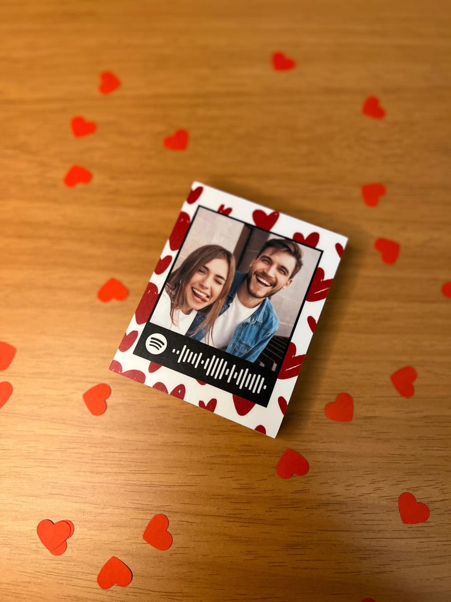 Arte Polaroid Namorados Canva com QR Code Spotify 3 Cartão branco com foto de casal sorrindo e código Spotify, sobre madeira com corações.