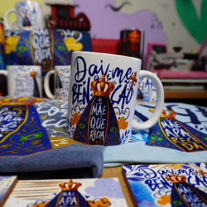 Caneca branca com arte digital religiosa, cercada por itens de devoção.