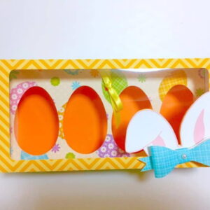 Caixa retangular amarela com estampa de ovos e orelhas de coelho decorativas.