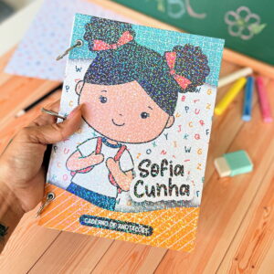 Caderno de anotações com capa brilhante, ilustrado com menina e letras, segurado por uma mão.