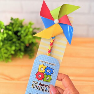 Mão segura cartão azul com catavento colorido, mensagem sobre amor e diferenças.
