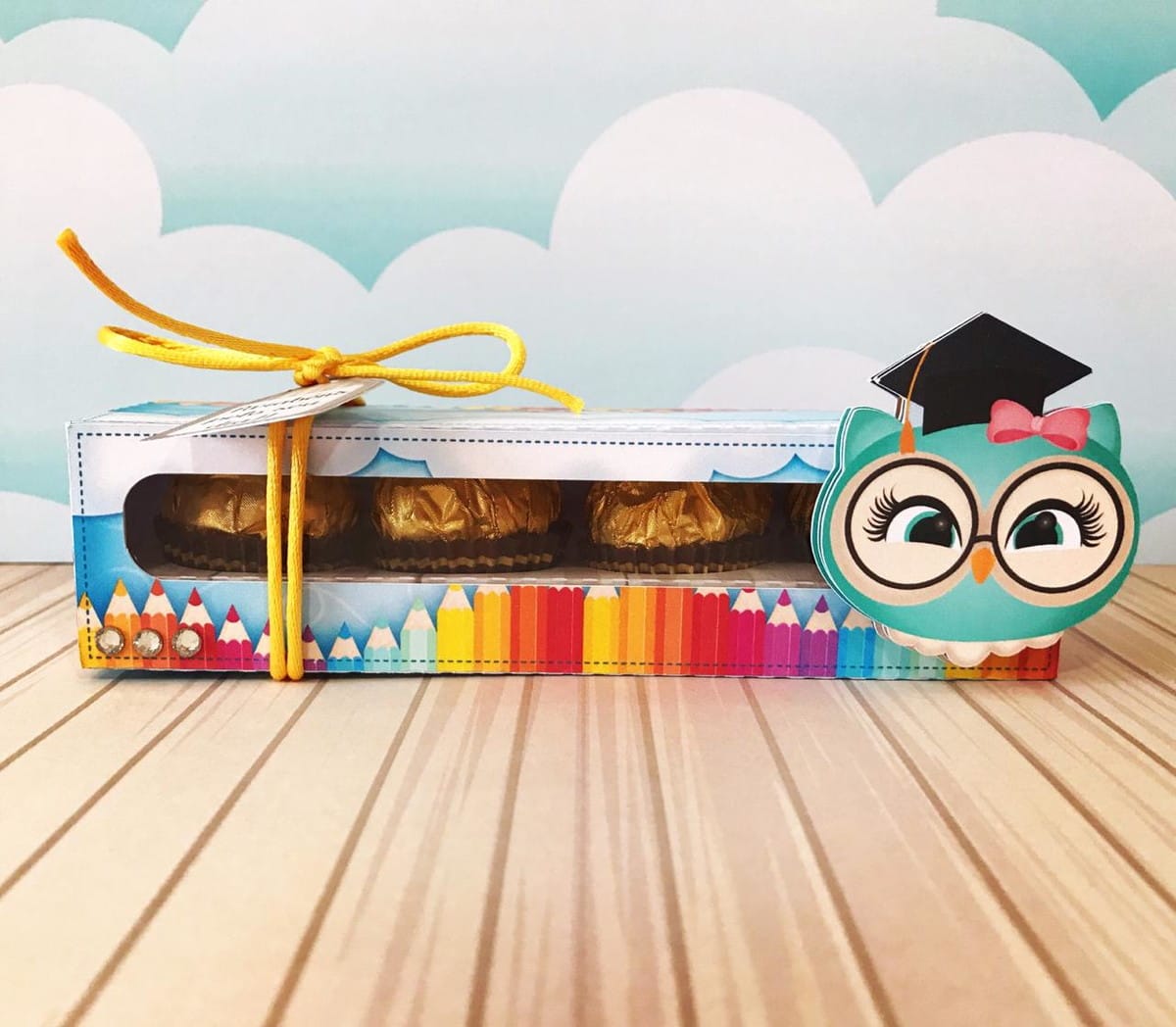 Kit Digital Dia dos Professores com Lembrancinhas Escolares 5 Caixa de presente decorada com coruja de formatura, lápis coloridos e doces embrulhados em dourado.