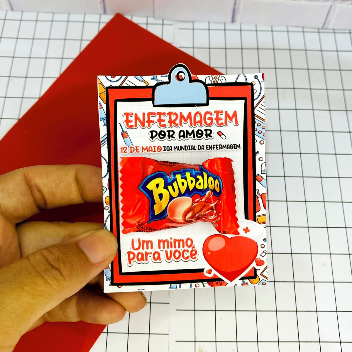 Kit Digital Enfermagem para Mimos e Lembrancinhas 3 Cartão temático "Enfermagem por Amor" com chiclete, segurado por uma mão.