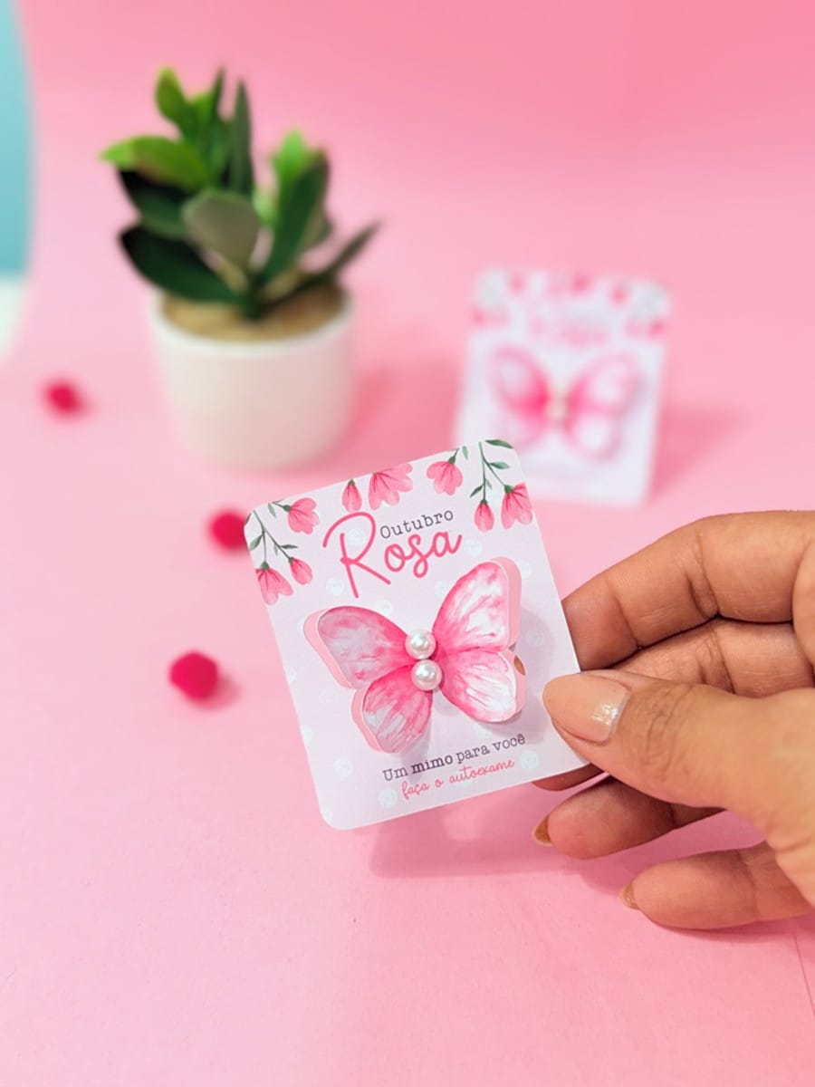 Kit Digital Outubro Rosa para Lembrancinhas e Mimos 3 Mão segurando cartão rosa com desenho de borboleta e texto "Outubro Rosa".