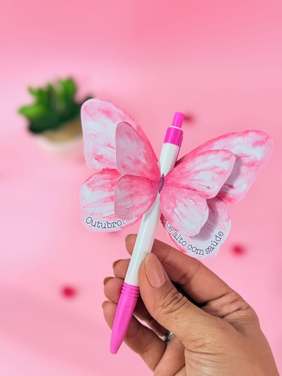 Kit Digital Outubro Rosa para Lembrancinhas e Mimos 1 Mão segurando caneta branca e rosa com asas de borboleta de papel.