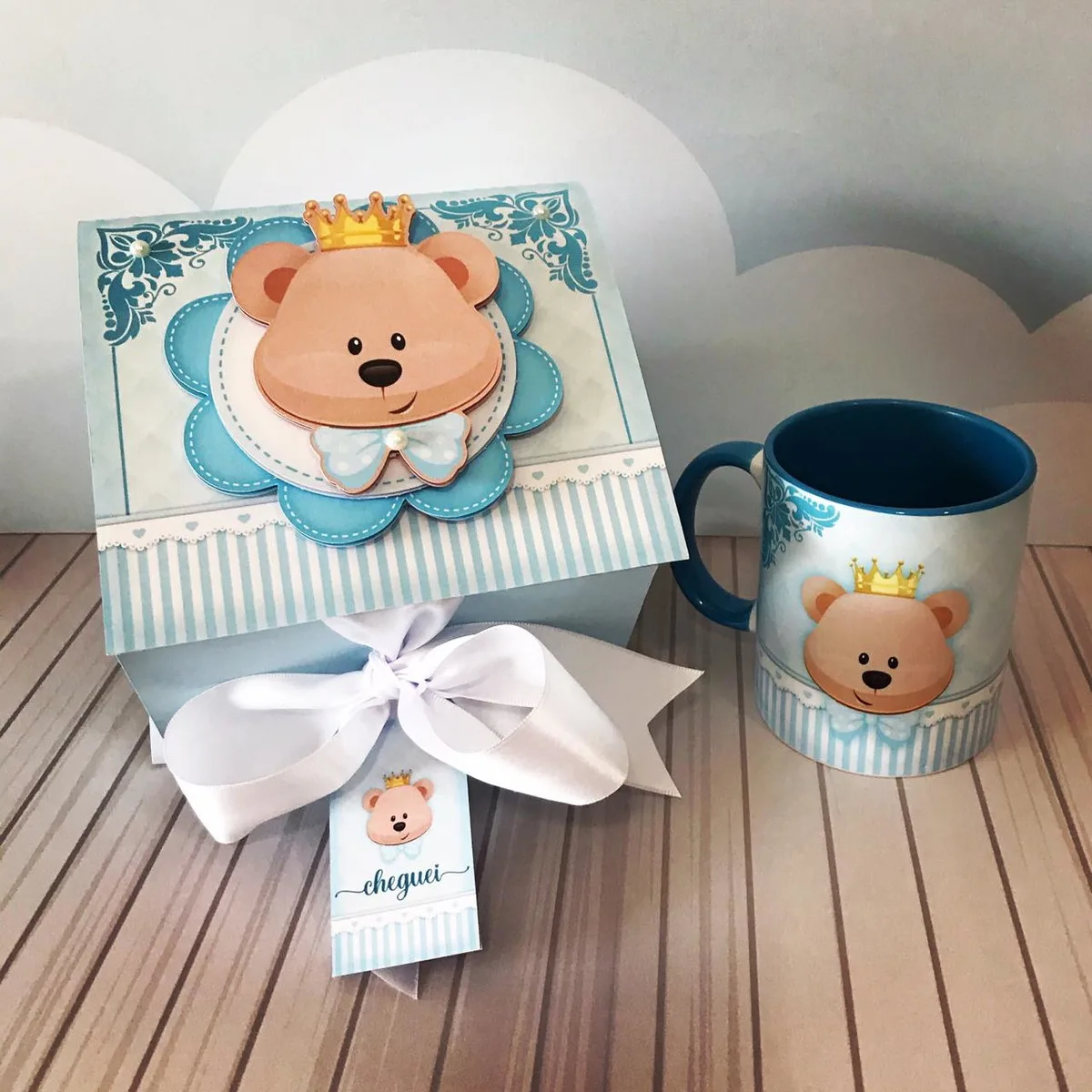 Caixa azul decorada com urso de coroa e caneca combinando sobre mesa listrada