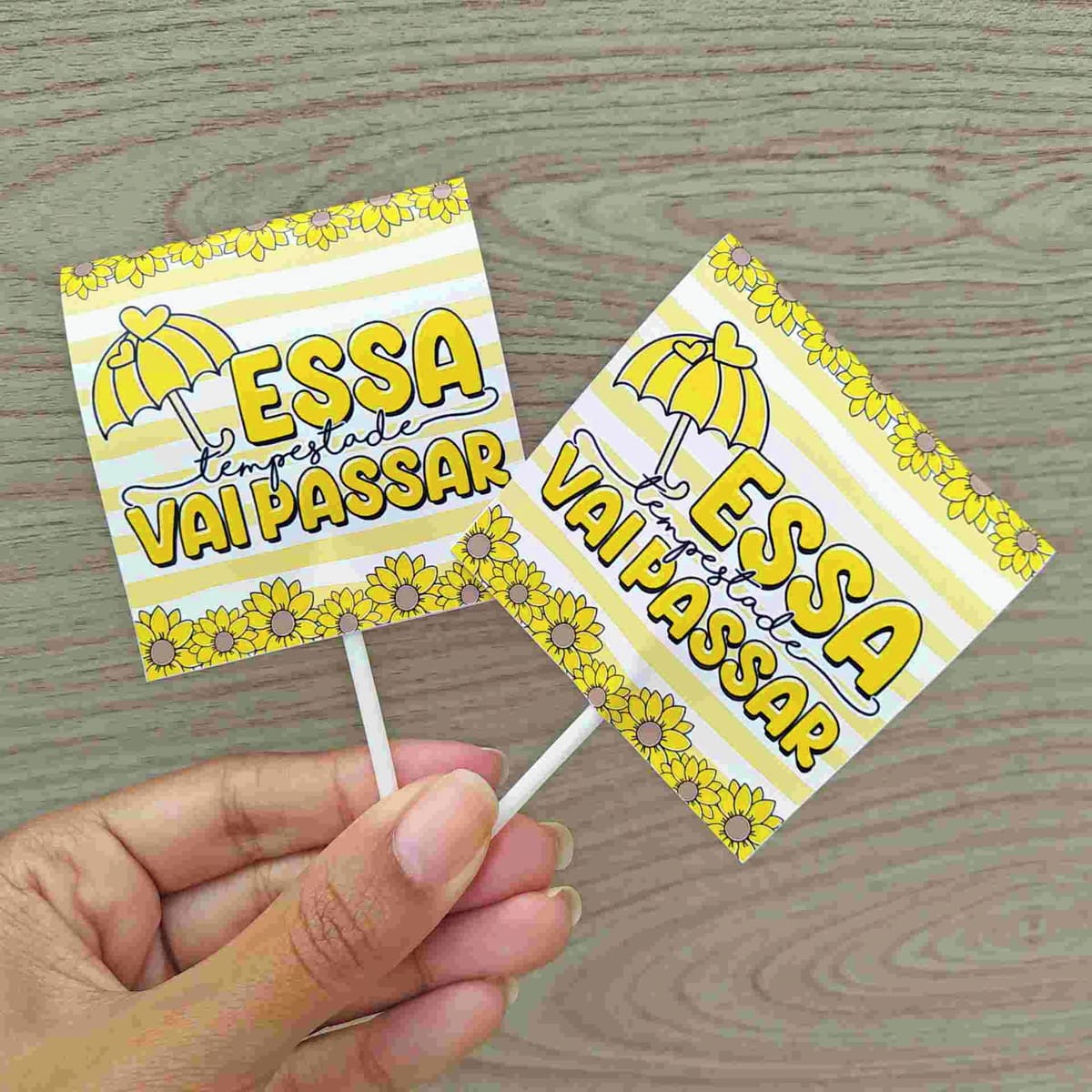 Kit Setembro Amarelo Digital com PNG para Imprimir 2 Mão segurando dois toppers amarelos com desenhos de guarda-chuva e girassóis sobre fundo de madeira.