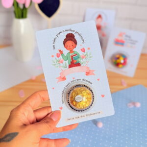 Mão segura cartão para professora com ilustração e bombom dourado.
