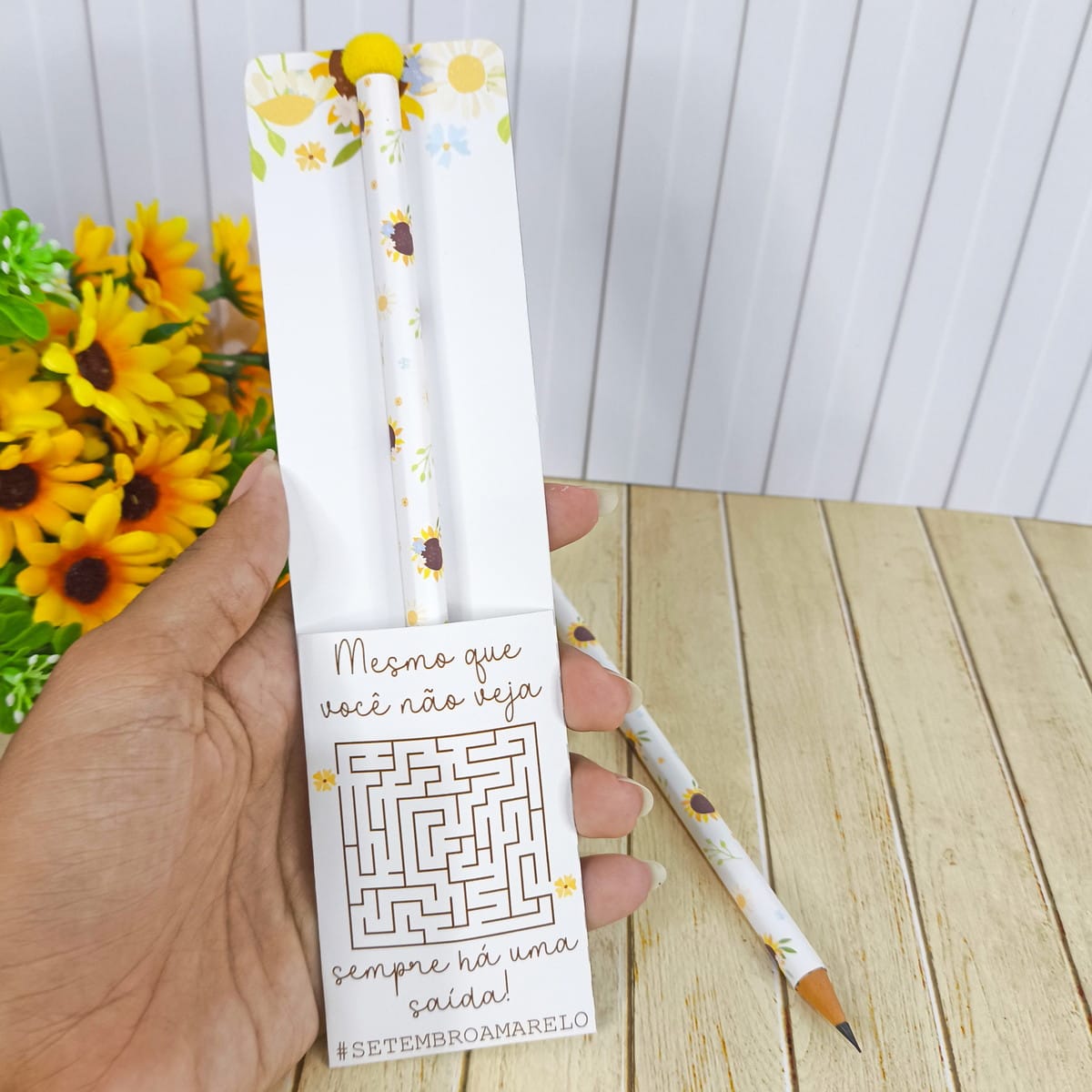 Kit Digital Setembro Amarelo Lembrancinhas em PNG 5 Mão segura cartão branco com dois lápis estampados, um labirinto e flores amarelas ao fundo.