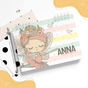 Caderno espiral colorido com ilustração de fada, caneta e clip sobre fundo branco.