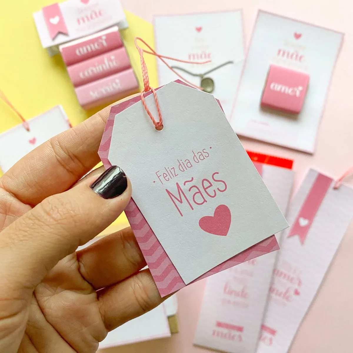 Mão segura etiqueta de papel com frase e coração ao lado de lembrancinhas rosas.