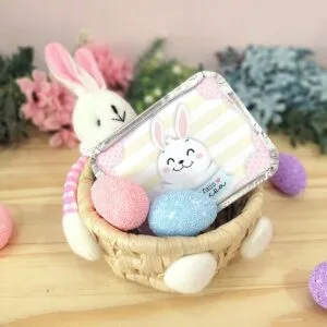 Cesta de vime com coelho de pelúcia, ovos coloridos e uma embalagem decorada.