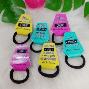 Porta Xuxinha Dia das Mães - Arquivo Digital 42 Cinco prendedores de cabelo pretos com cartões coloridos estampados dispostos sobre superfície felpuda branca.